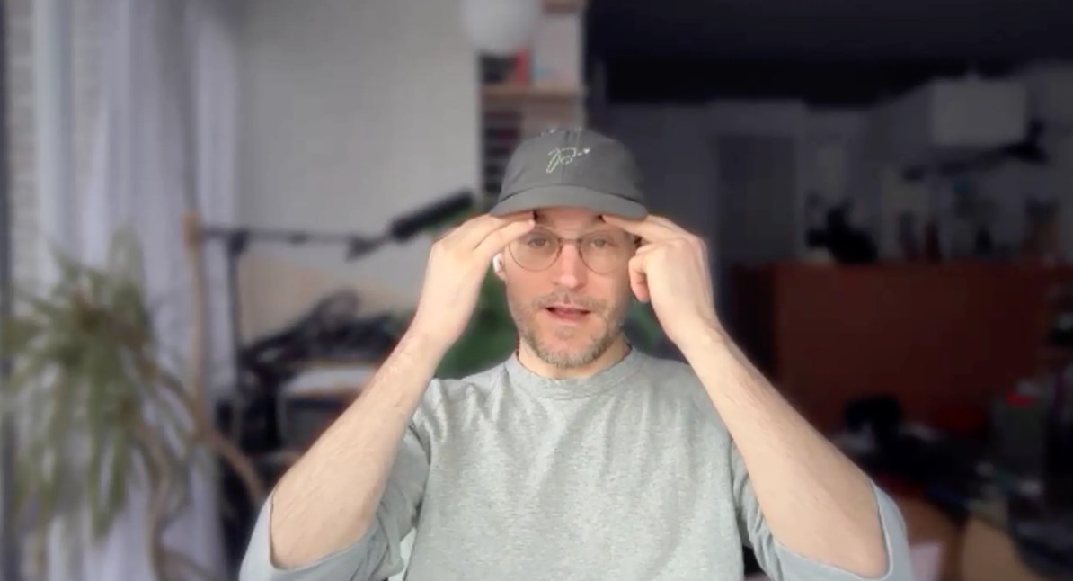 Author Craig Mod on a zoom call, wearing a dark grey baseball cap and light grey long sleeve shirt, his fingers to his head in a gesture of mind explodeyness.